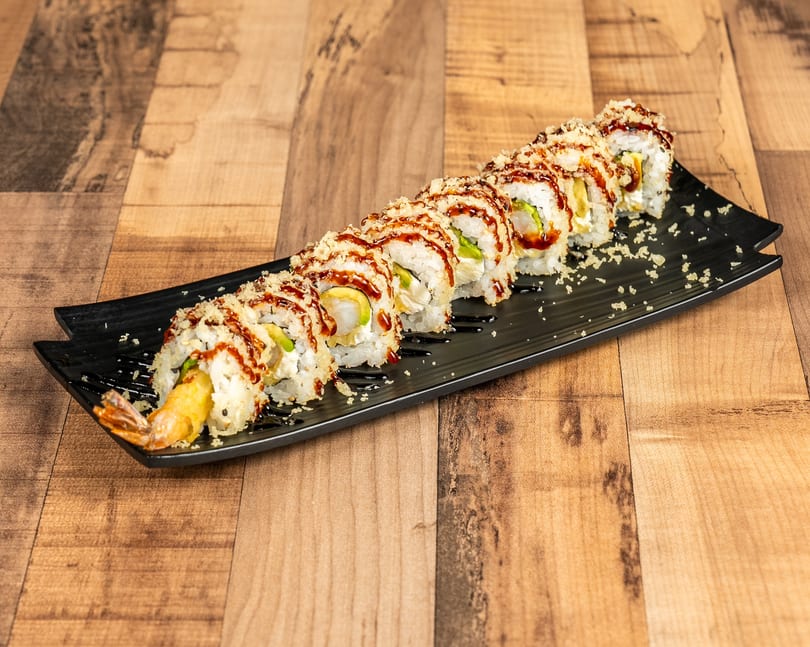 A long black plate holds a row of sushi rolls with shrimp tempura, avocado, and sauce.