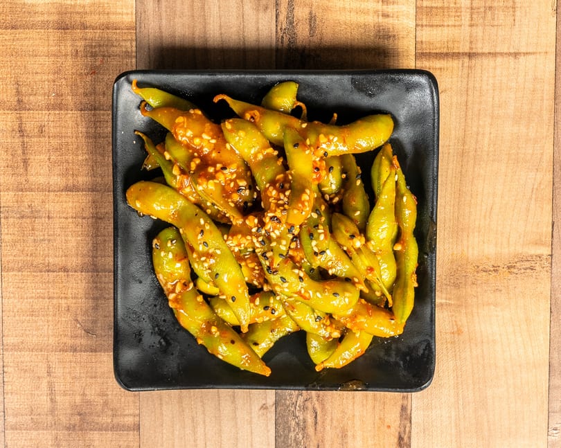 Spicy edamame with sesame seeds in a black rectangular dish on a wooden table.