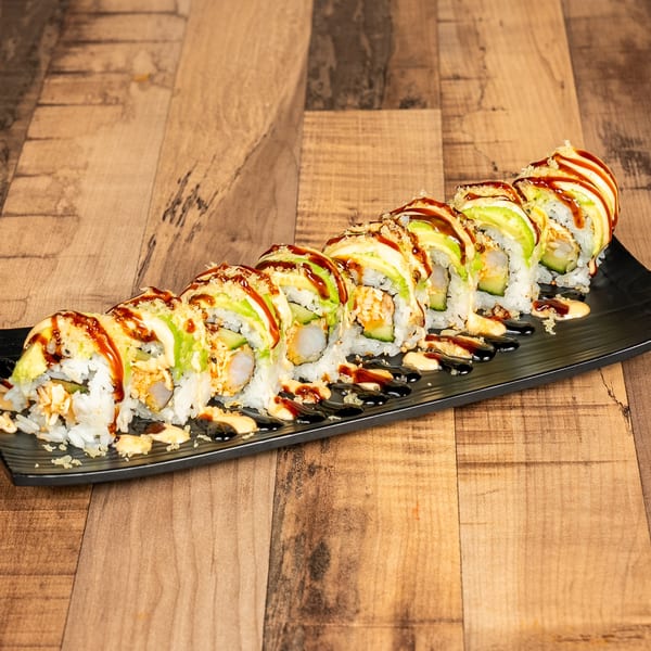 A long black plate holds a row of sushi rolls topped with avocado, sauces, and crunchy bits.