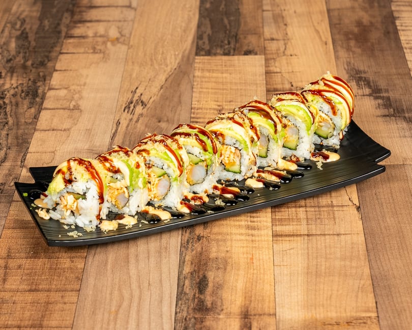 A long black plate holds a row of sushi rolls topped with avocado, sauces, and crunchy bits.