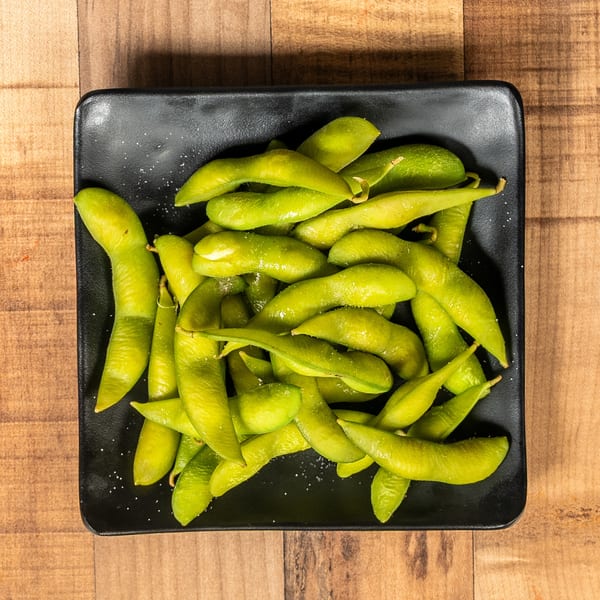 A black rectangular plate filled with bright green edamame pods, lightly sprinkled with salt, on a wooden table.