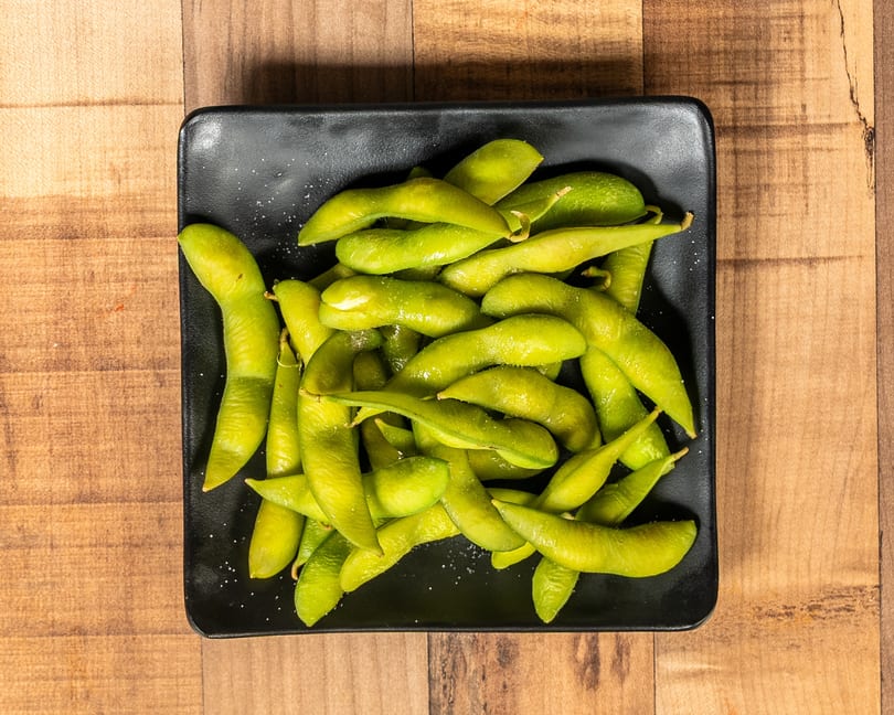 A black rectangular plate filled with bright green edamame pods, lightly sprinkled with salt, on a wooden table.
