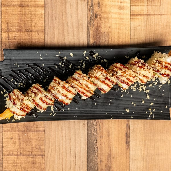 Tempura shrimp sushi roll with dark sauce drizzle and crispy flakes on a black rectangular plate.